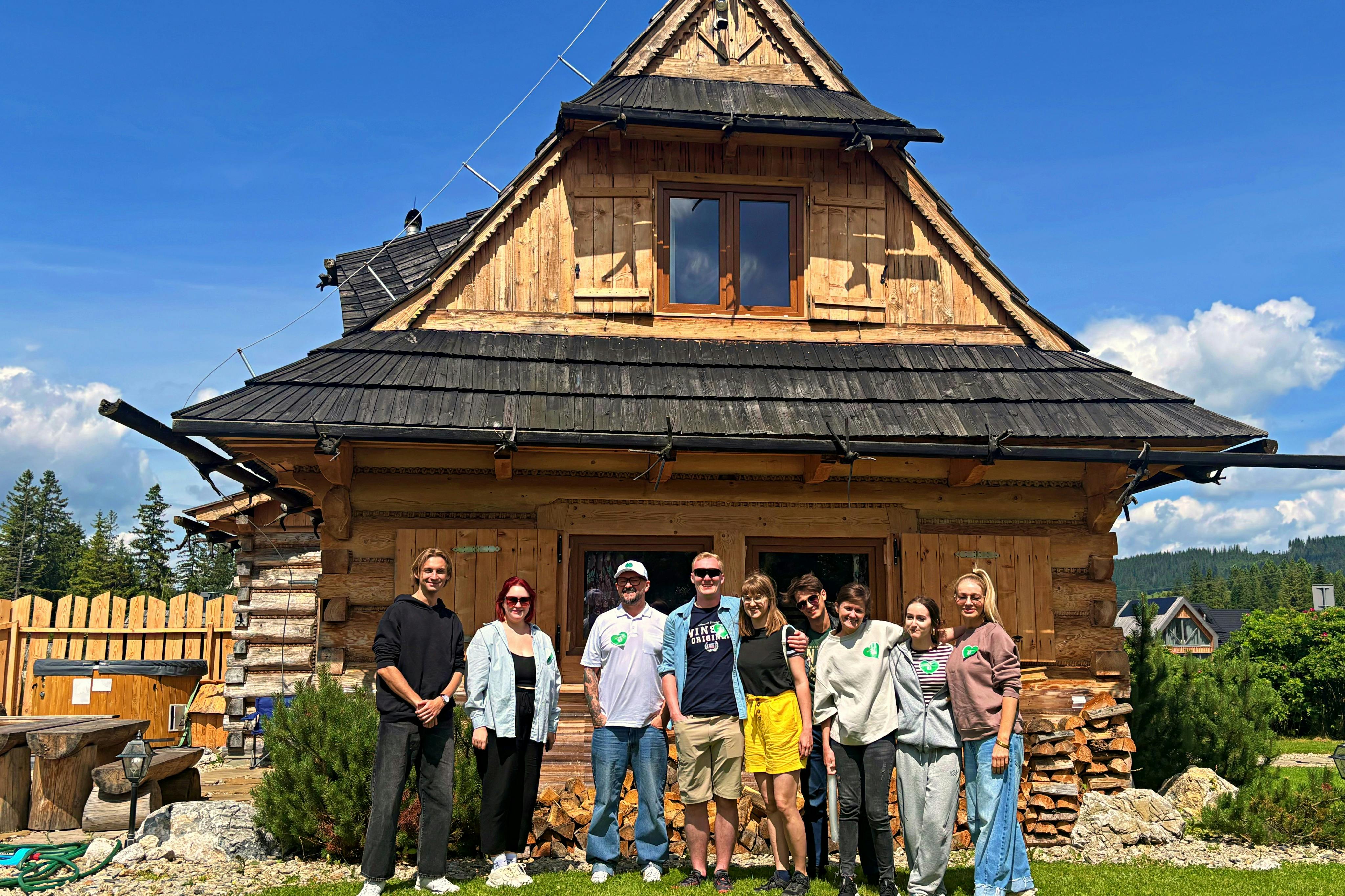 木々と青い空を背景に、急な屋根の素朴な木造の家の前に8人が立っています。