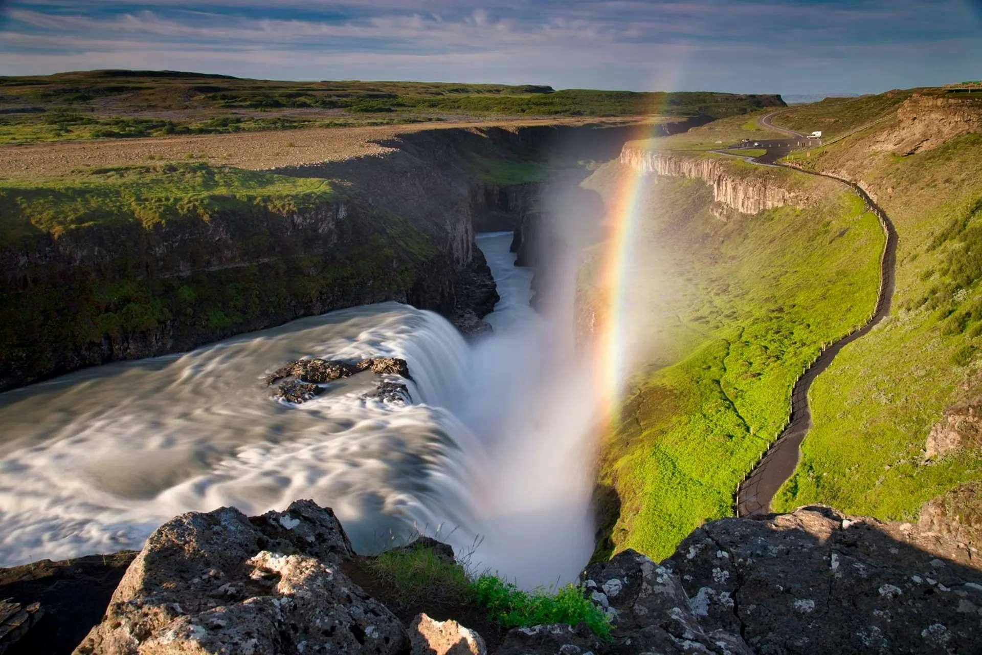 Powerful waterfall cascading into a canyon with a vibrant rainbow arching over it, surrounded by lush green landscape.