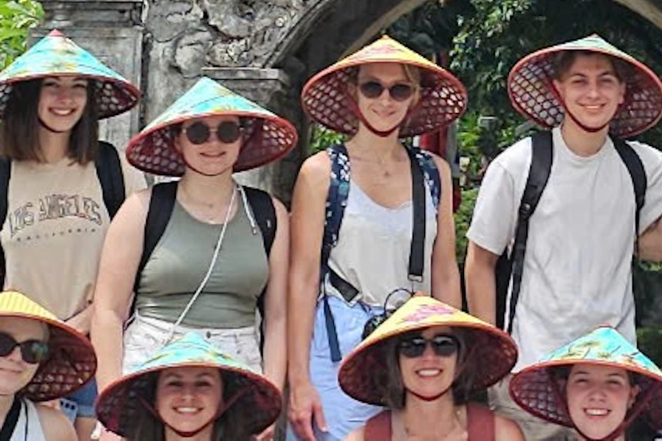 A group of people wearing conical hats pose in front of an ornate stone gate surrounded by greenery.