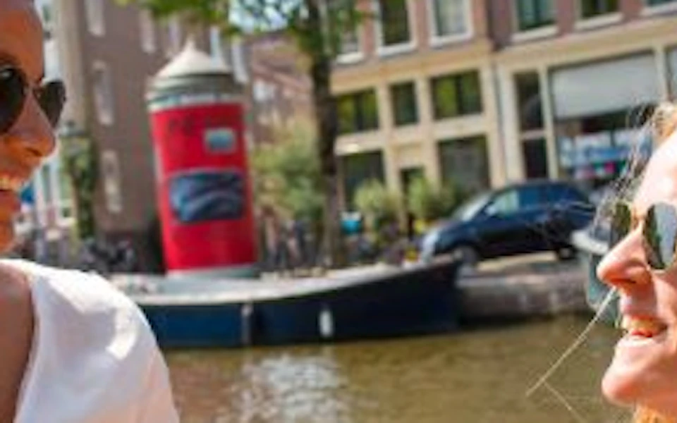 Two women smiling and holding sandwiches on a boat. Background shows a canal, buildings, and a red buoy.