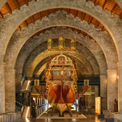 Ett utsmyckat, historiskt skepp som visas i en välvd stenbyggnad med träbjälkar och belysning på gångvägarna.