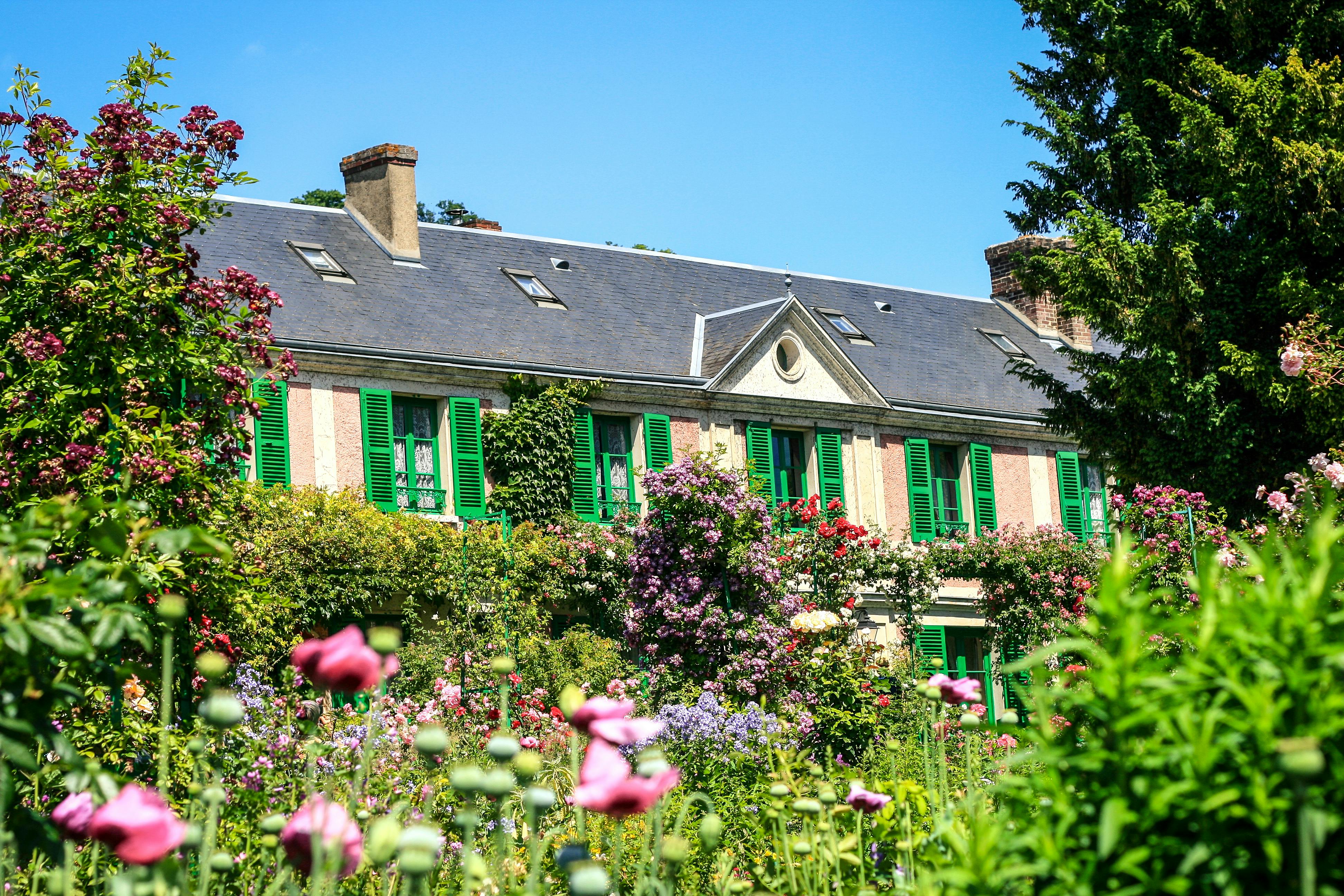Ένα διώροφο σπίτι με πράσινα παντζούρια, περιτριγυρισμένο από καταπράσινους κήπους και πολύχρωμα λουλούδια κάτω από έναν καταγάλανο ουρανό.
