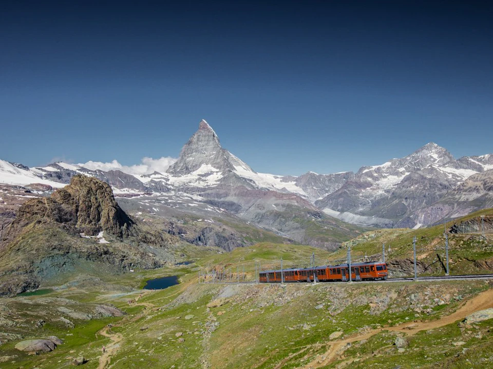 A red train travels through a mountainous landscape with the snow-capped Matterhorn peak in the background under a clear blue sky.