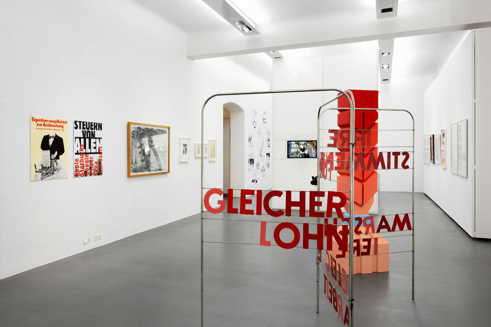 Umělecká galerie s různými zarámovanými uměleckými díly na bílých stěnách a kovovou konstrukcí s červeným textem „GLEICHER LOHN“.