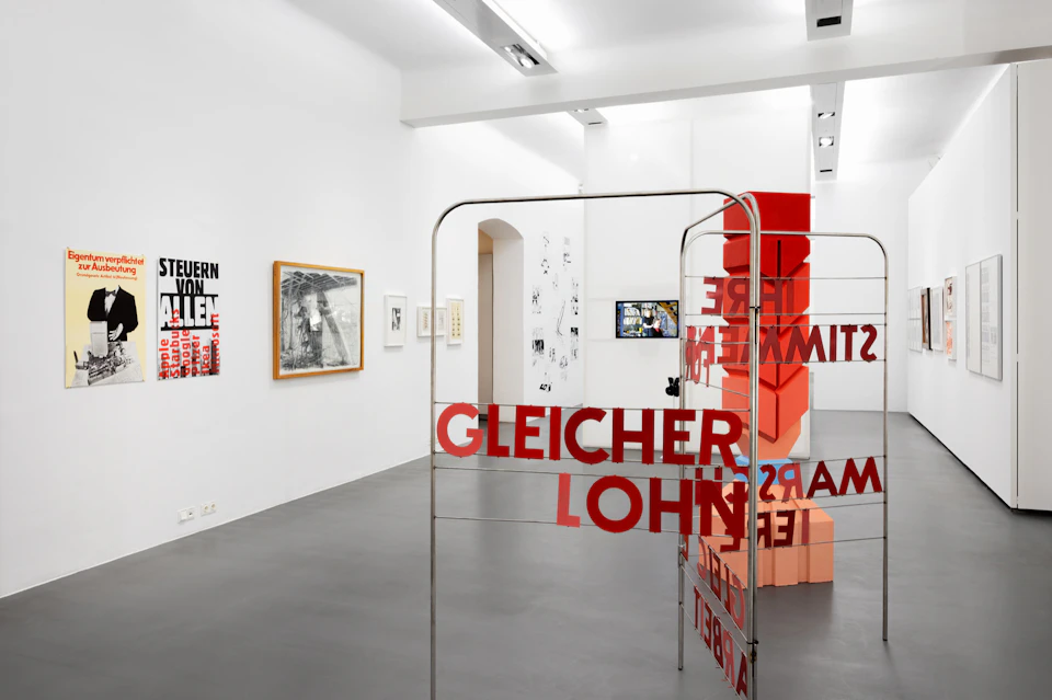 An art gallery featuring various framed artworks on white walls and a metal structure with red text "GLEICHER LOHN."