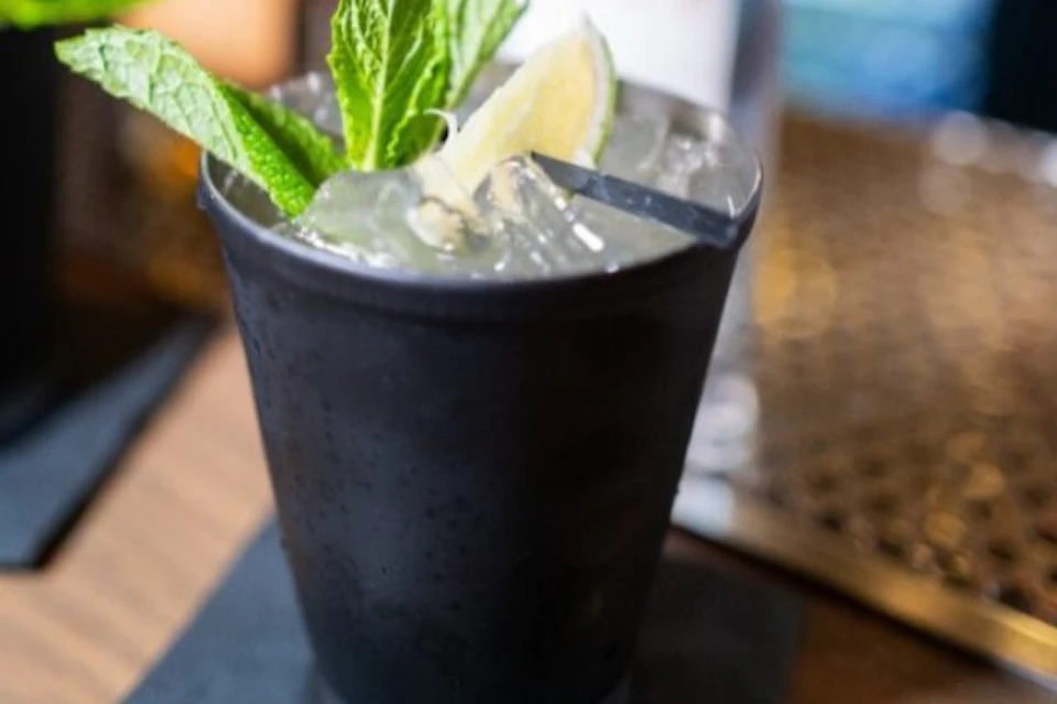 A black cup with a mint sprig, lime wedge, and straw in a clear beverage, sitting on a napkin on a wooden surface.