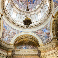 Ornate ceiling of a church with detailed frescoes, a large central dome, and an intricate chandelier hanging in the center.