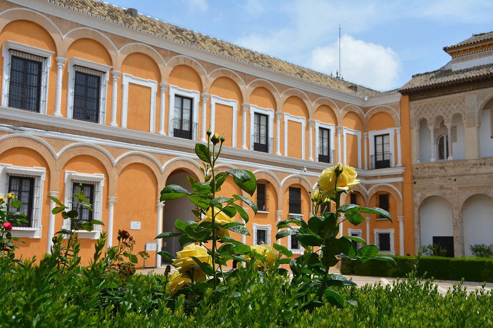 Yellow roses in a garden in front of a building with orange walls, white arches, and a tiled roof under a partly cloudy sky.