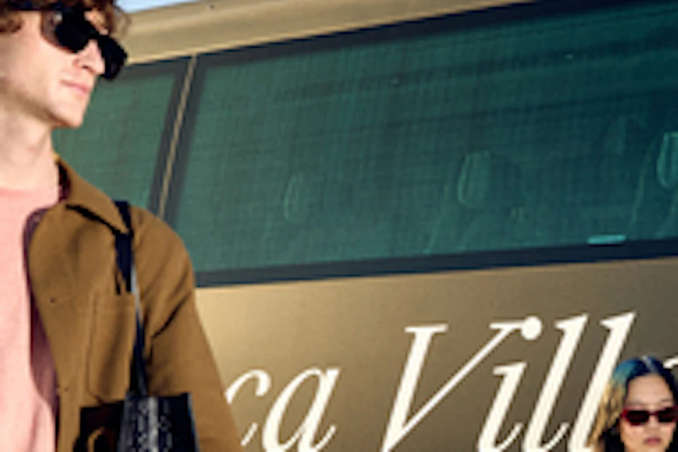 A man and woman with shopping bags stand near a bus labeled "La Roca Village to La Roca Village" on a sunny day.