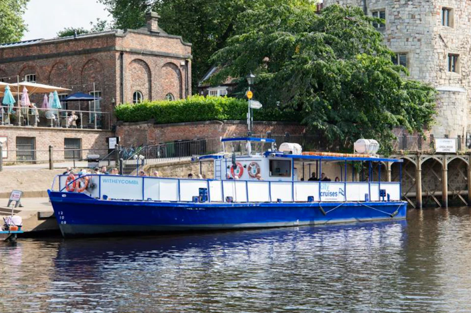 A blue and white riverboat named "Witheycombe" docked beside a brick building near lush green trees and a stone structure.