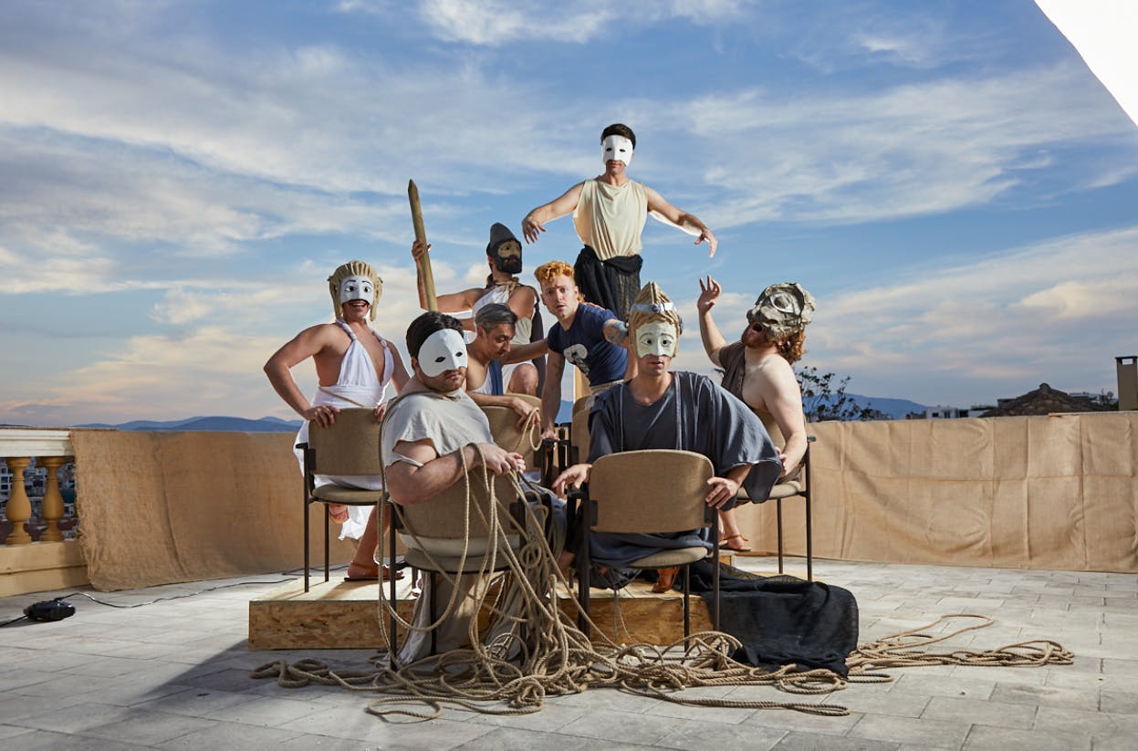 一群戴着白色面具和各种服装的人在户外布景上摆姿势，蓝天下摆着椅子和绳子。