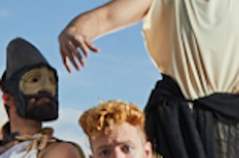 A group of people wearing white masks and various costumes pose on an outdoor set with chairs and ropes under a blue sky.