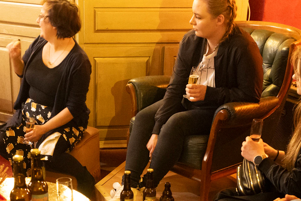 People sitting in a cozy room, chatting and drinking, with bottles and glasses on tables.