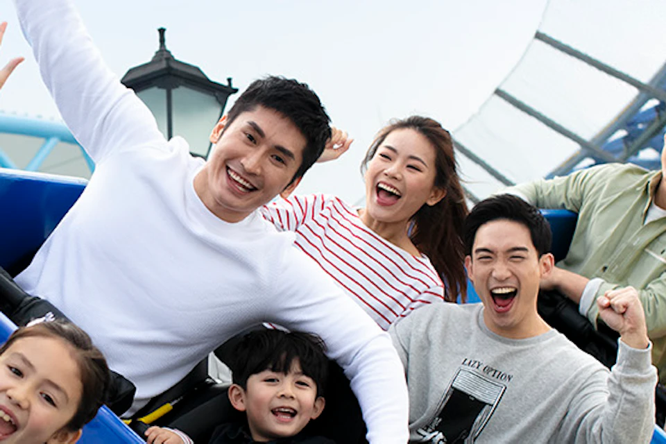 A group of people smiling and raising their hands while riding a roller coaster on a sunny day.