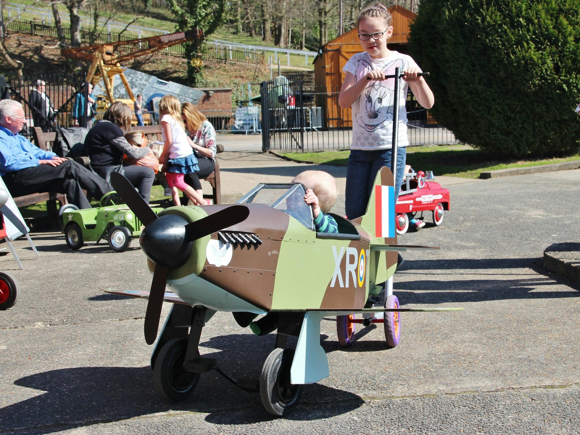 Una nena empeny un nen en un petit avió de joguina en una zona asfaltada, amb persones i un cotxe de joguina al fons.