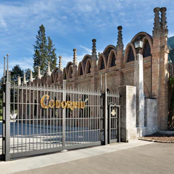 Utsmyckad smidesjärnsport med ordet "Codorníu" i guld, framför en gammal tegelbyggnad med valv och träd i bakgrunden.