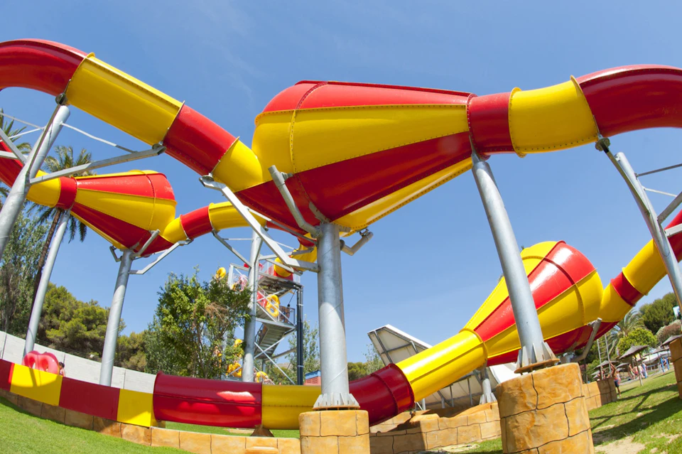 A brightly colored, intertwining red and yellow water slide structure in an outdoor amusement park under a clear blue sky.