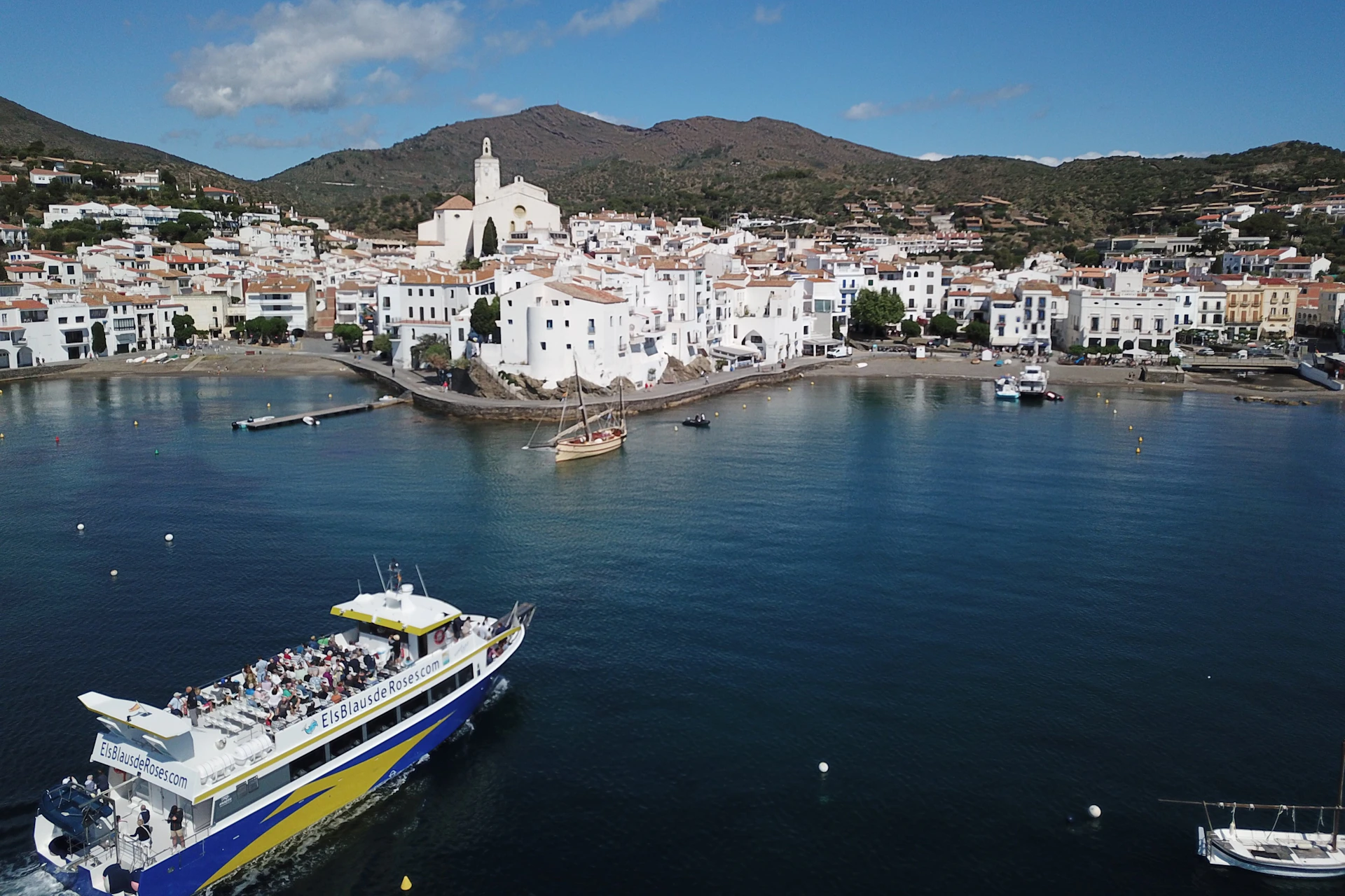 A coastal town with white buildings, a church and a boat in the foreground, set against calm blue water beneath a partly cloudy sky.