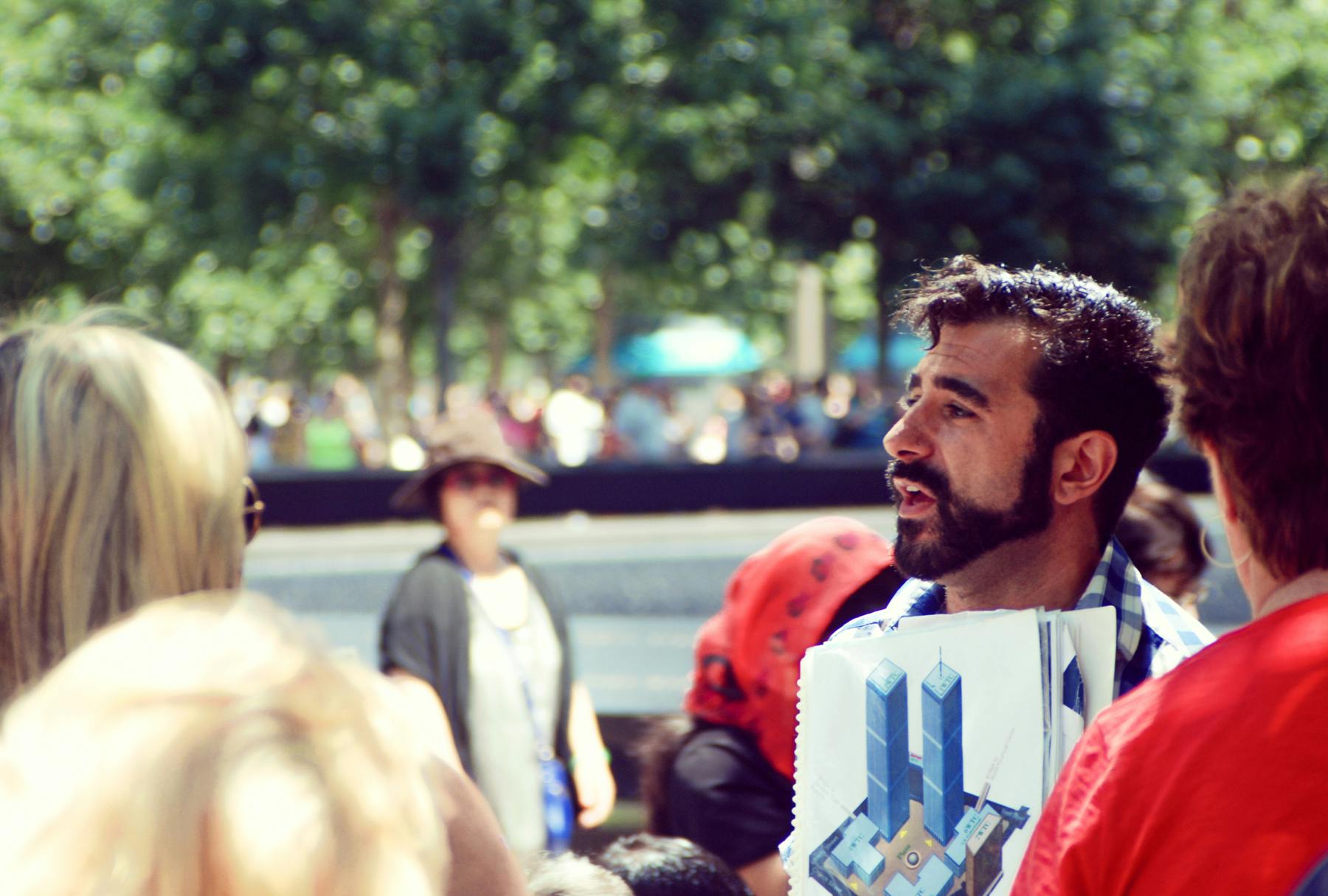 Um homem de barba segurando uma imagem impressa das Torres Gémeas, no meio de uma multidão ao ar livre, com árvores desfocadas ao fundo.