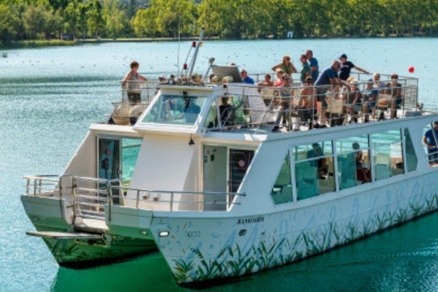 Un vaixell de passeig blanc en un llac amb gent a la coberta superior. Al fons hi ha arbres i una costa verda.