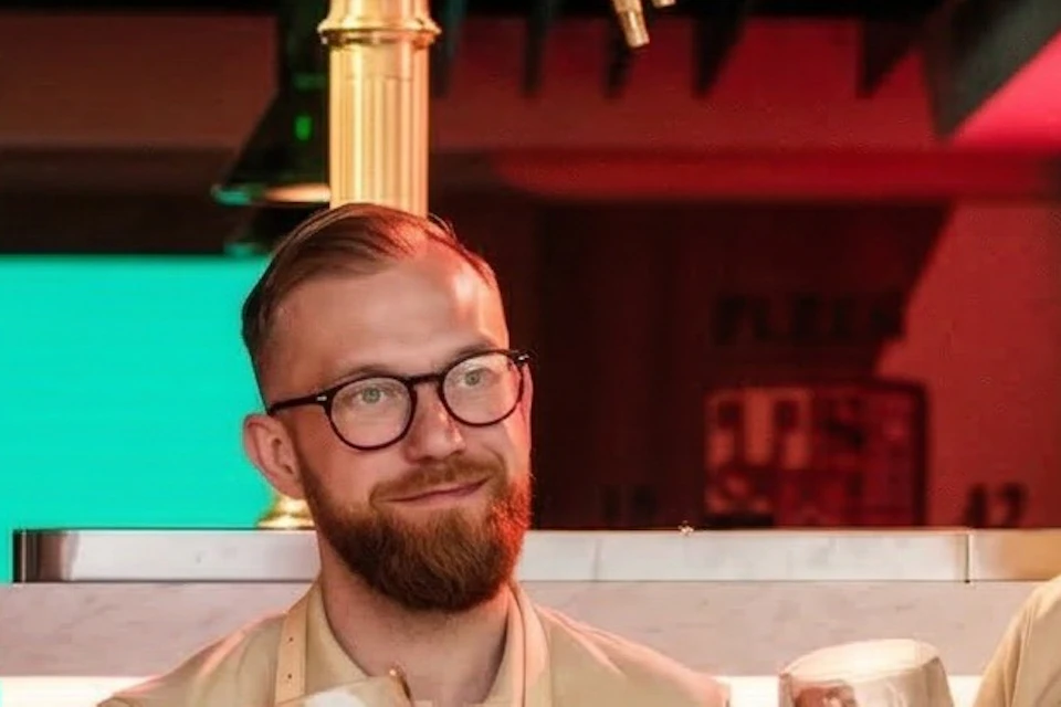 Four bartenders wearing aprons stand behind a bar, smiling and holding mugs of beer in a brightly lit setting.
