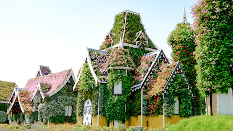 Whimsical houses covered in colorful flowers and greenery, with pointed roofs and arched windows, set against a bright sky.