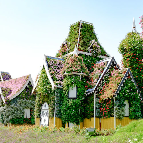 Whimsical houses covered in colorful flowers and greenery, with pointed roofs and arched windows, set against a bright sky.