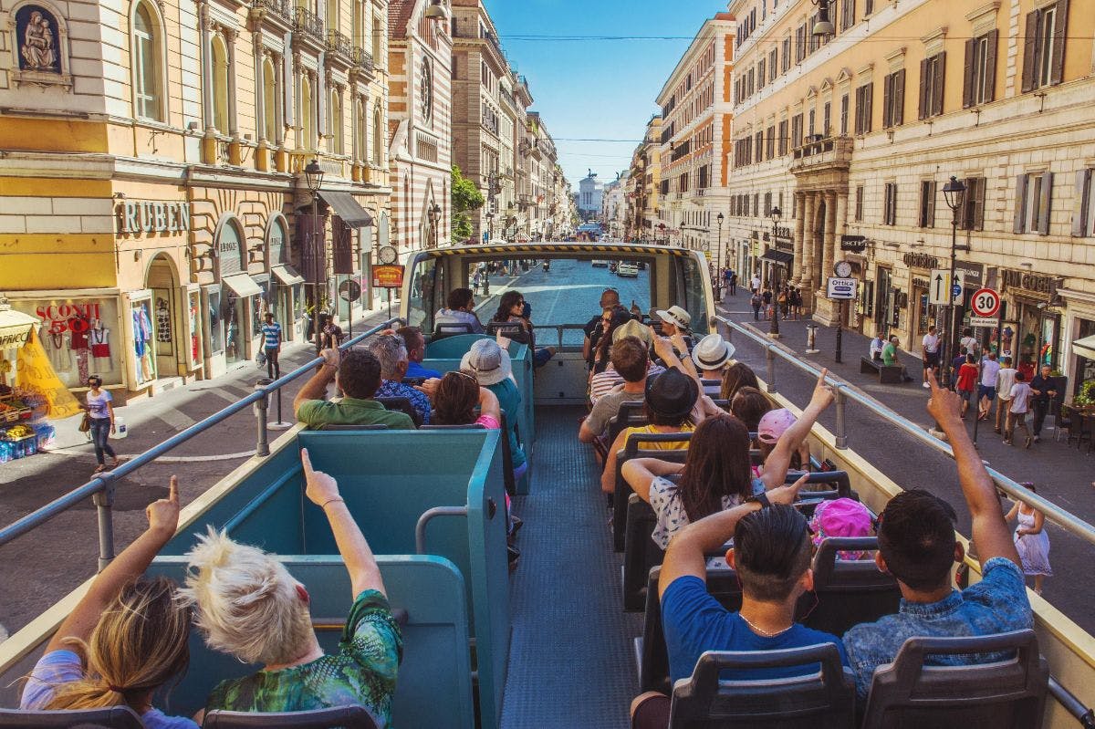People on an open-top tour bus in a busy European city street with historic buildings and shops on either side.