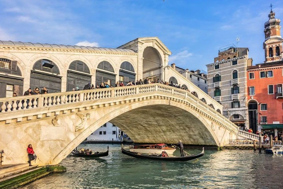 Stone arch bridge over a canal with gondolas below and people walking on top, set against a clear blue sky.