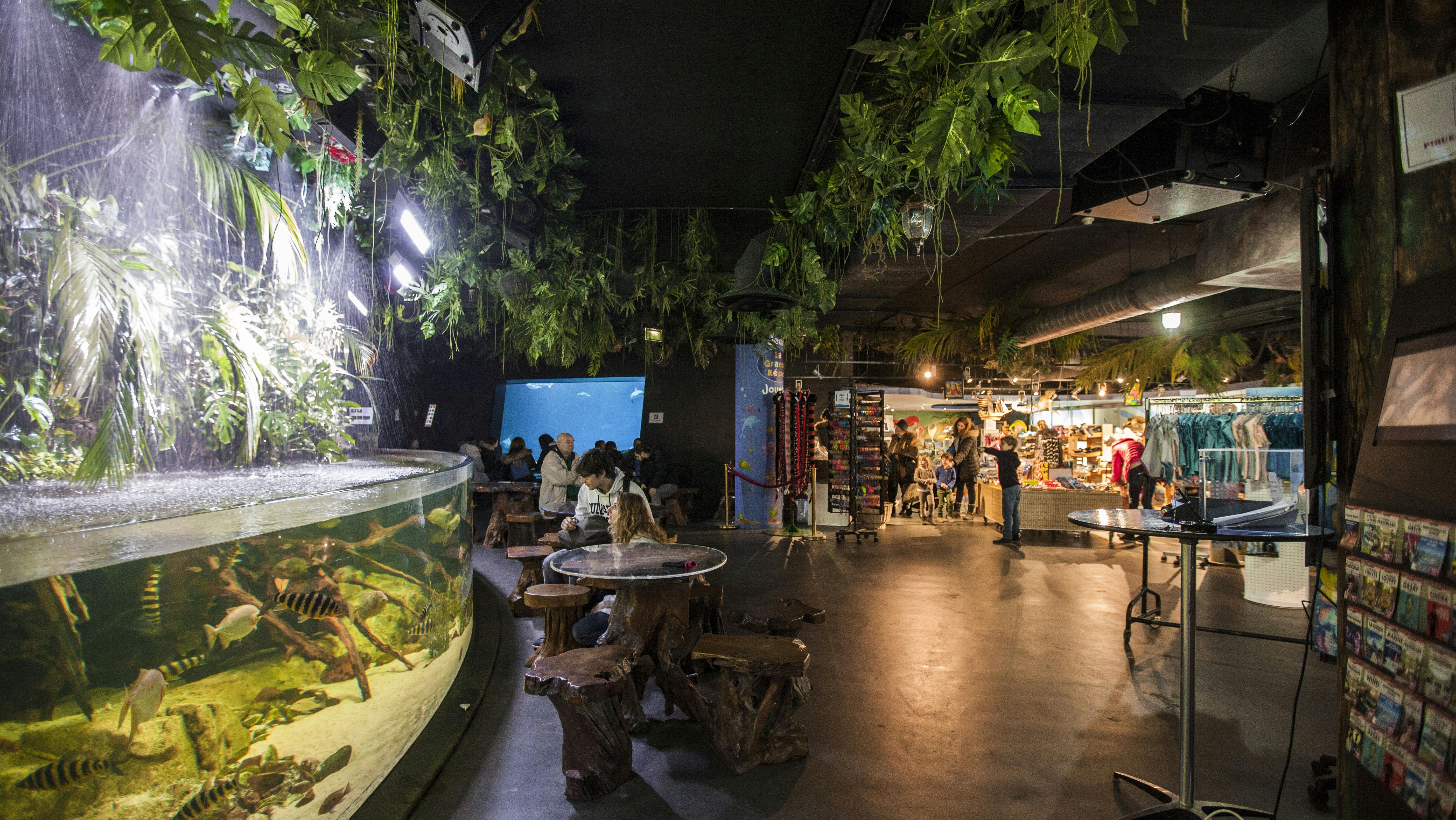 Cena de interior com pessoas sentadas em mesas rústicas perto de um grande aquário, rodeadas por plantas suspensas e uma loja de recordações próxima.
