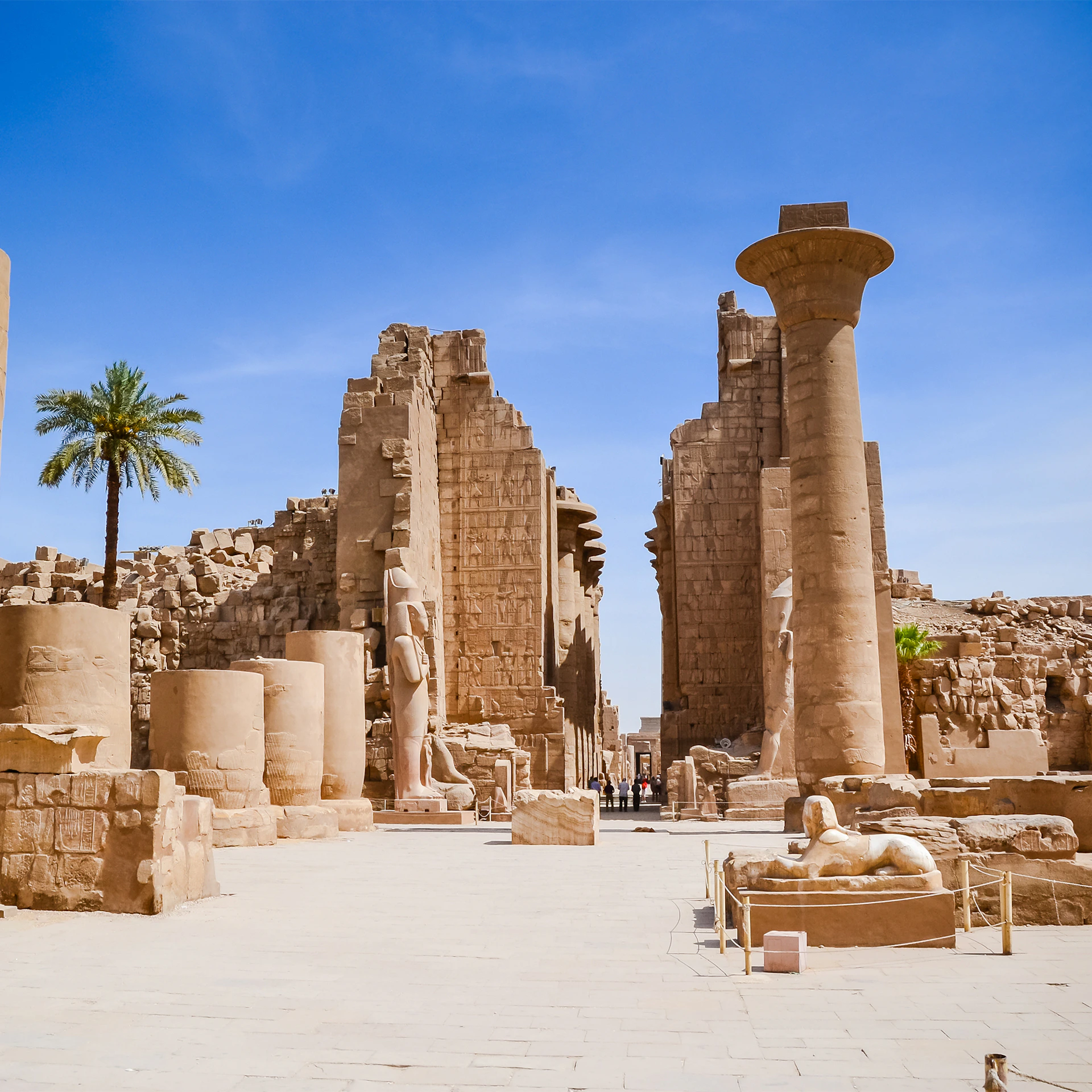 Ancient ruins with tall stone columns, statues, a palm tree, and remnants of walls under a clear blue sky. People in the distance.