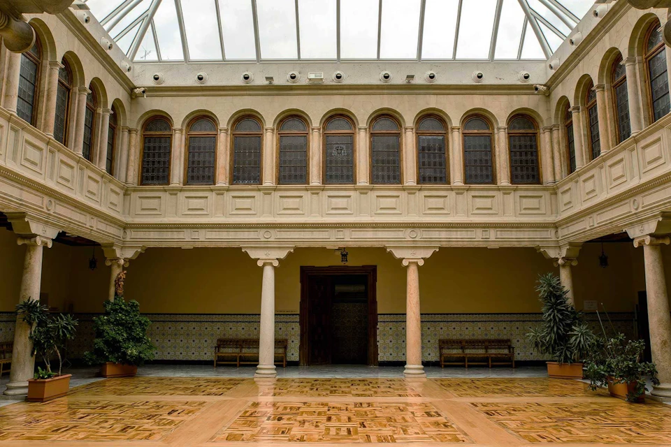 A spacious atrium with a skylight roof, arched windows on the upper level, and tiled walls and floor with plants on the sides.