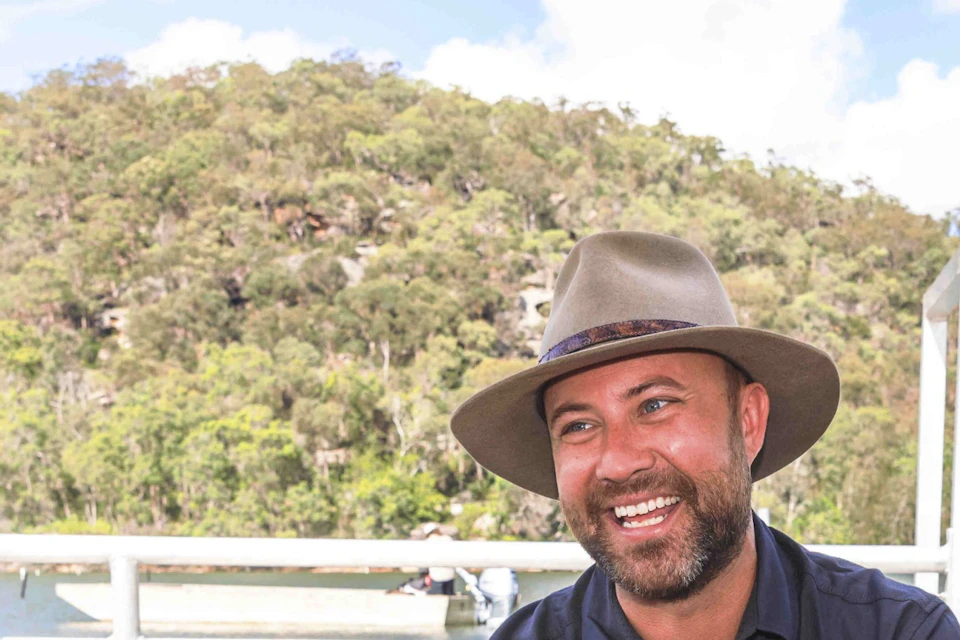 A group of people on a boat engage with a man presenting oysters, surrounded by scenic water and greenery.