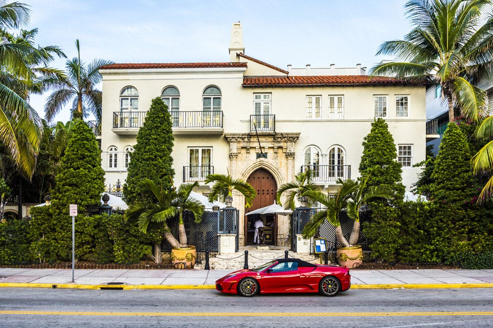 A large, upscale white building with an arched entrance and palm trees in front. A red sports car is parked on the street.