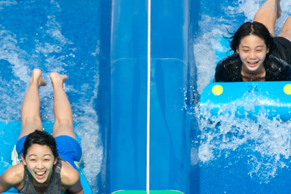 Four people sliding down parallel green and blue water slides, smiling and holding onto mats. Trees are in the background.