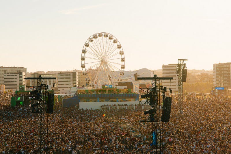A large crowd at an outdoor event with a Ferris wheel and stage structures under a sunny sky.