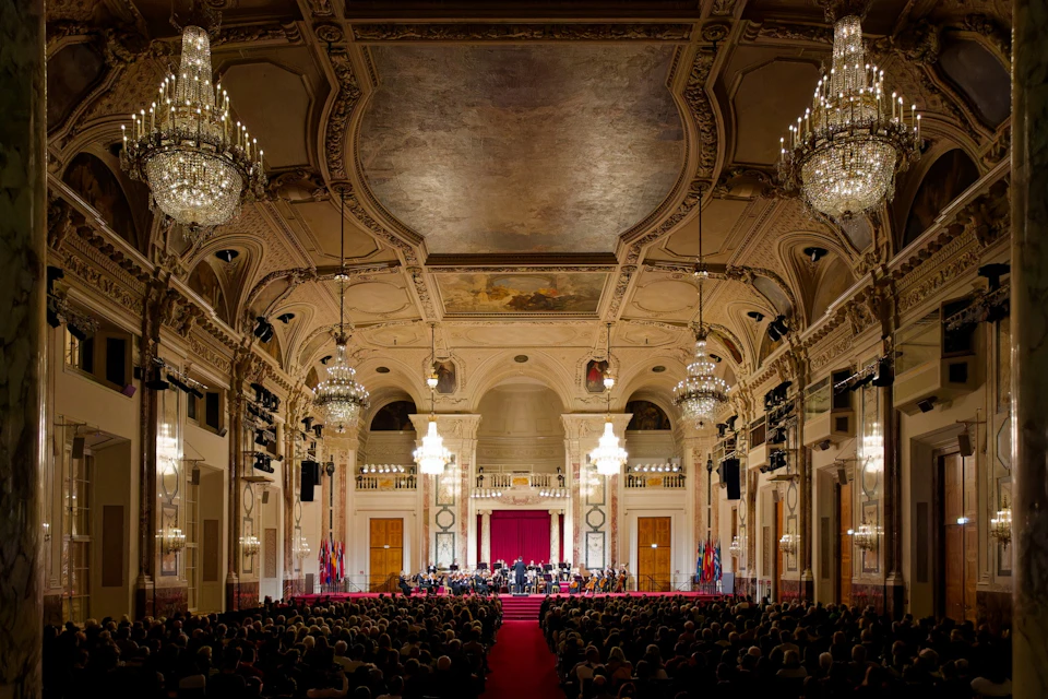 An orchestral performance in a grand hall with ornate chandeliers and a domed ceiling, attended by a large audience.