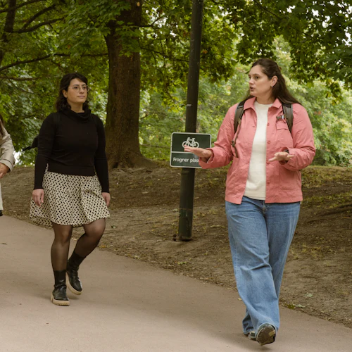 Fyra personer som går på en stig i en park, omgiven av träd, med en skylt med texten "Frogner plass" synlig.