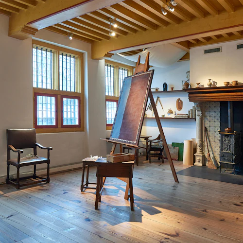 Konstnärsateljé med staffli, stol, träbord och öppen spis. Hyllor med keramik och böcker syns i bakgrunden.