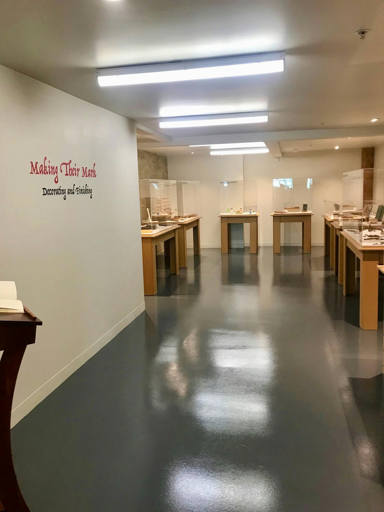 Museum tentoonstelling met boeken op tafels, glazen vitrines en een bord aan de muur met de tekst "Making Their Mark: Decorating and Embellishing".