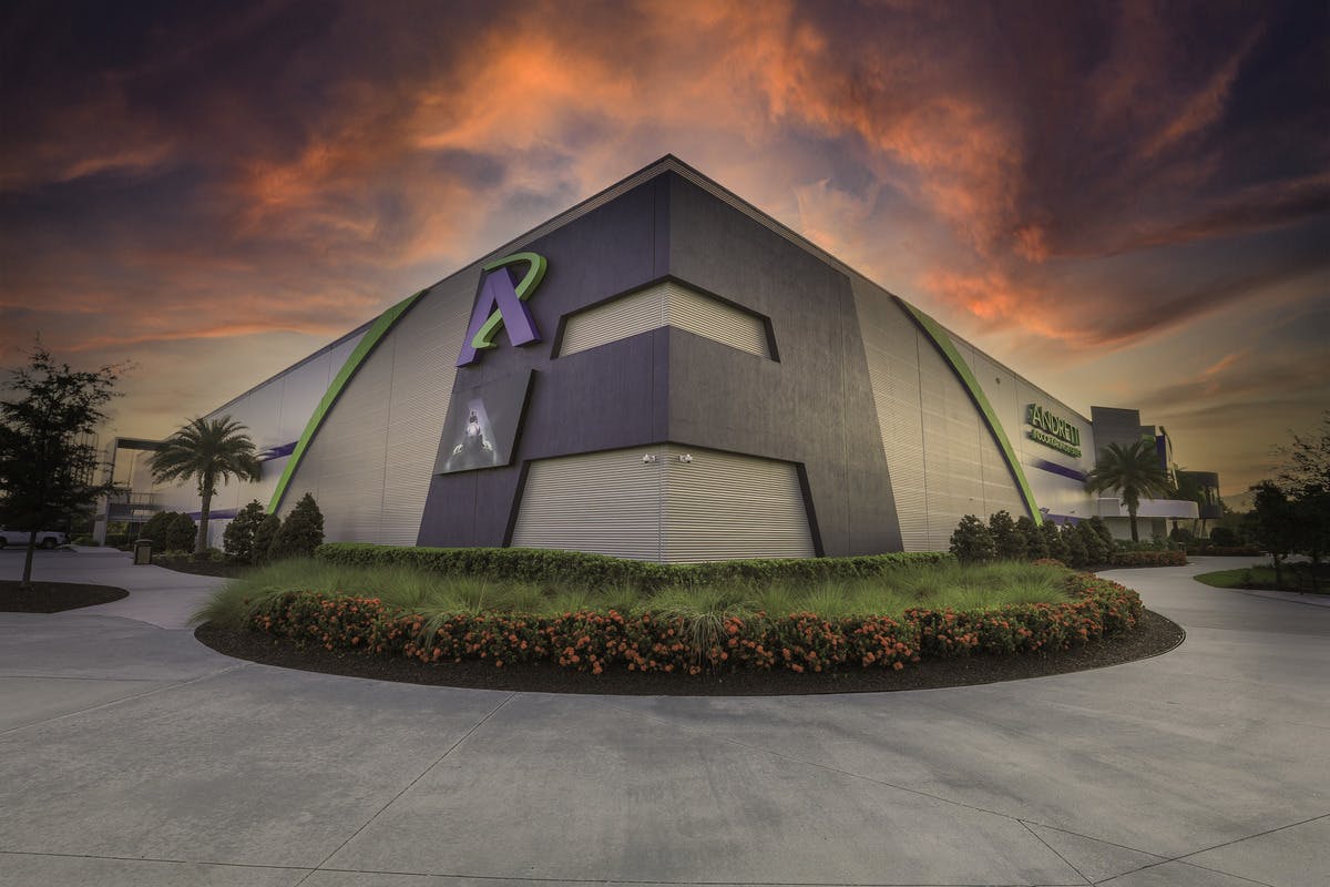 A large, modern building with green accents under a dramatic, orange and purple sky, surrounded by landscaped bushes.