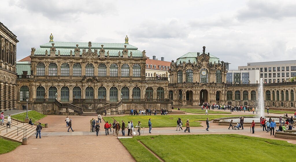Visitors at a historic palace courtyard with ornate architecture, a fountain, and manicured lawns under a cloudy sky.