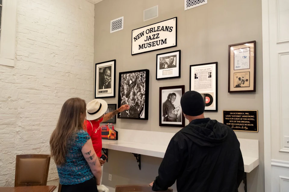 Three people examine framed jazz memorabilia on a museum wall, with a sign reading "New Orleans Jazz Museum" above them.
