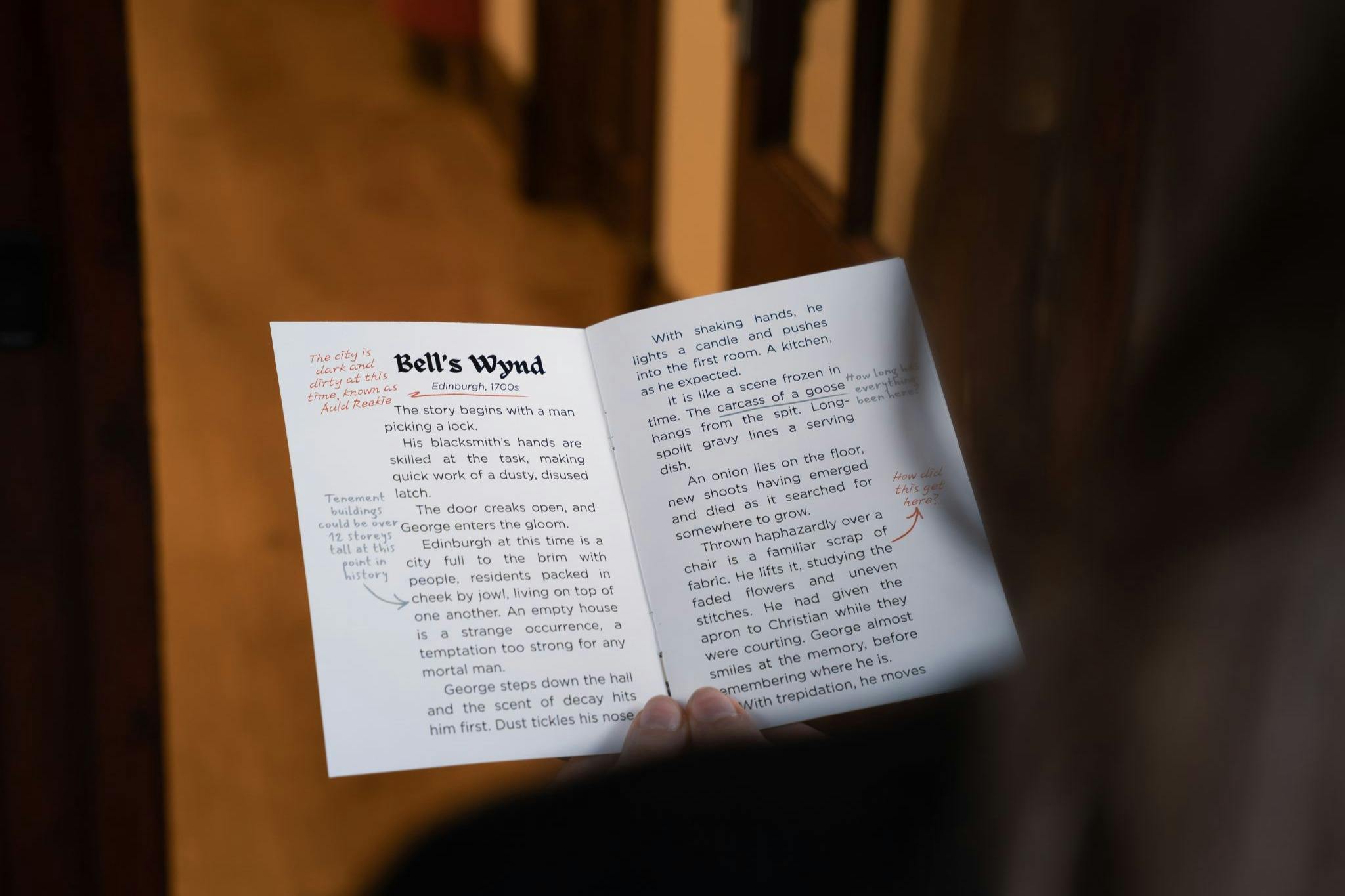 Una persona sostiene un folleto abierto titulado "Bell's Wynd" con texto impreso y notas manuscritas en tinta roja en las páginas.