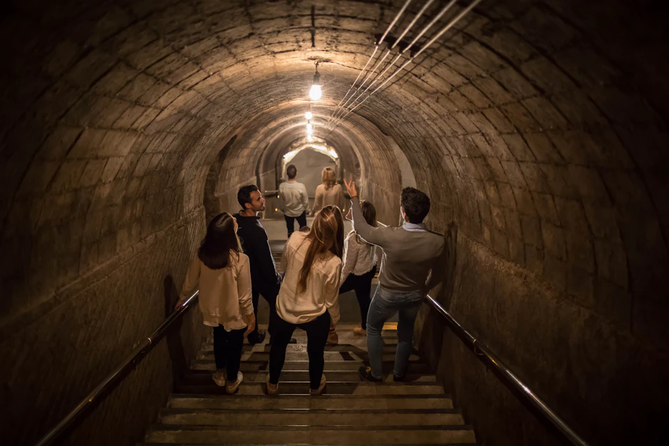 A group of people descends down a dimly lit, arched brick tunnel with staircases and handrails.