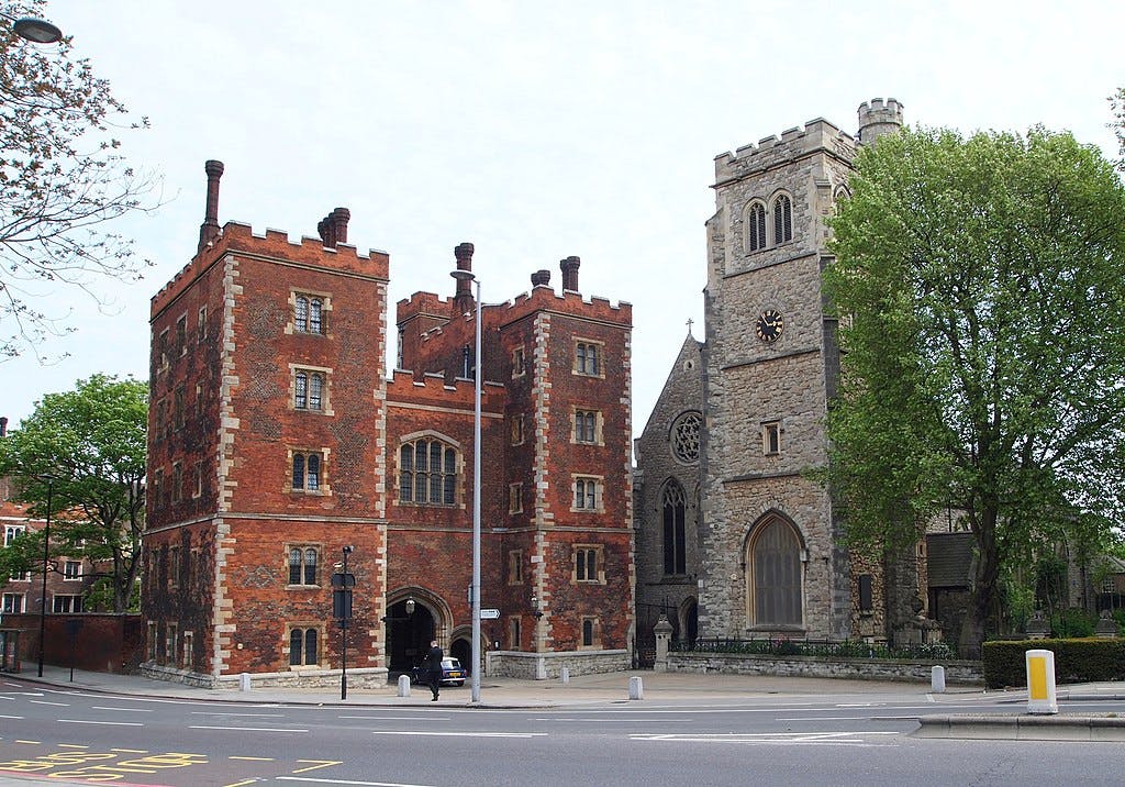 一栋带有城垛的红砖建筑矗立在一座石头教堂旁边，教堂设有钟楼，毗邻道路和绿色树木。