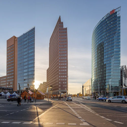 Tre moderna höghus i ett stadsområde i solnedgången, med bilar och fotgängare på gatan nedanför.