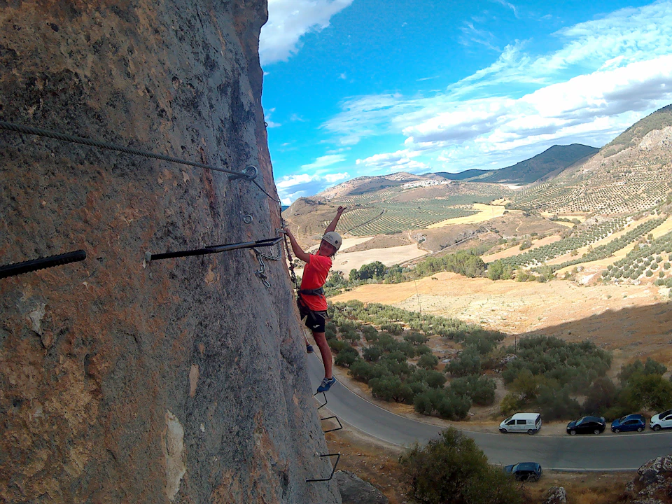 A climber in a red shirt scales a steep rock face with metal rungs, overlooking a scenic landscape of rolling hills and a winding road.