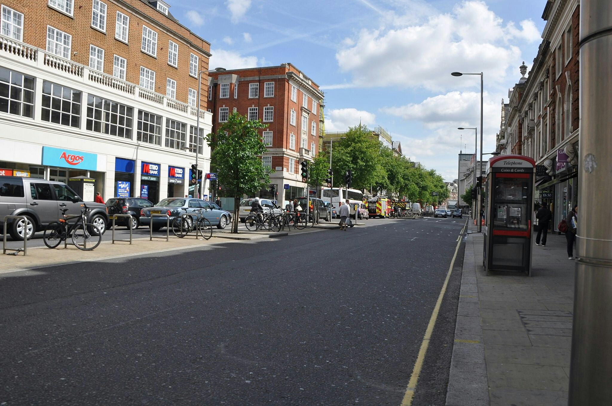Rue avec des voitures et des vélos garés, une cabine téléphonique, des magasins et des gens qui marchent. Bâtiments en briques et verdure sous un ciel bleu.