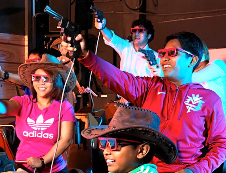 People wearing 3D glasses seated on motion simulator machines, holding guns and appearing to be engaged in an interactive ride.